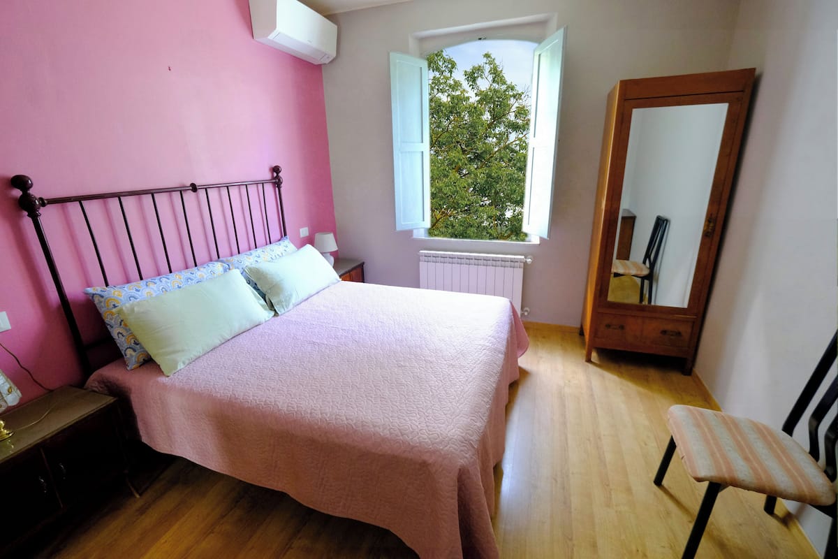 Une chambre à coucher comprenant un lit, une table de chevet, une chaise, une fenêtre donnant sur des arbres et un miroir.