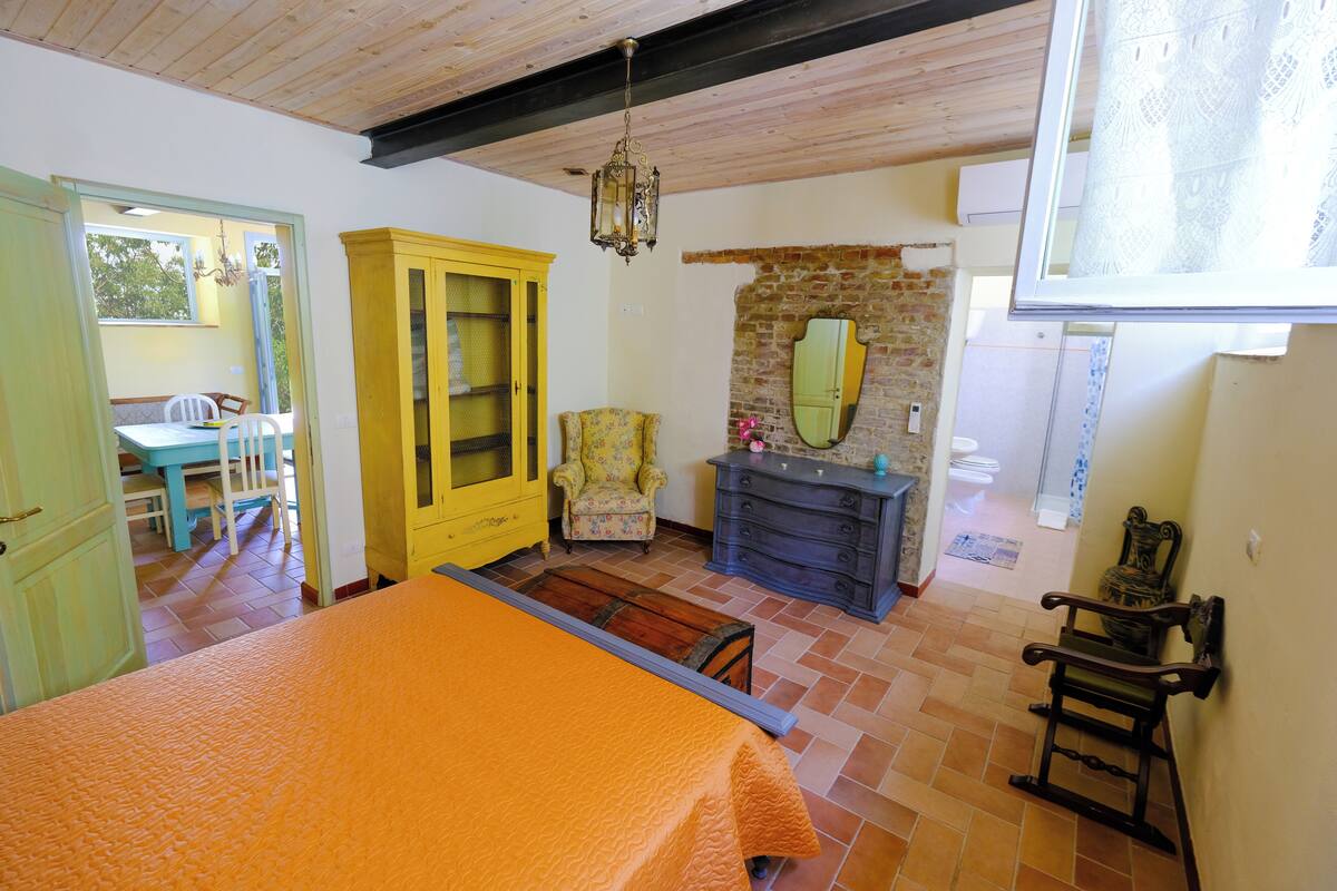 Une pièce avec un lit, un plafond en bois, une armoire jaune, un fauteuil à motifs floraux, une commode et un miroir sur un mur en briques.