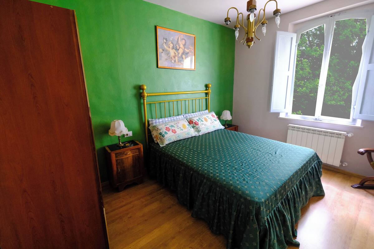 Une chambre avec un mur vert, un cadre de lit en bois, une couvre-lit verte, une table de chevet avec une lampe et une fenêtre donnant sur des arbres.