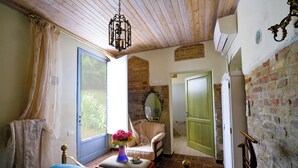 Un dormitorio con cama, una silla, una mesa con un florero y una ventana con cortinas, además de una puerta que da a otra habitación.