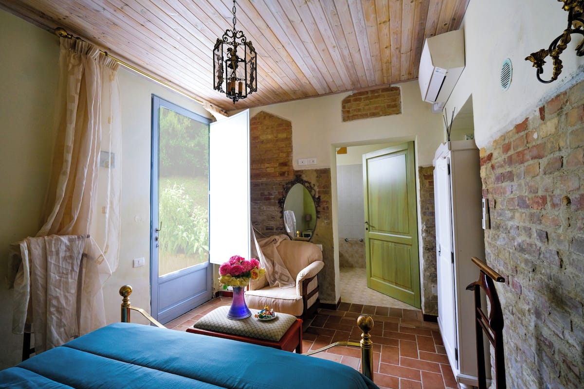 Une chambre à coucher comprenant un lit, une chaise, une table avec un vase de fleurs, une fenêtre avec des rideaux et une porte donnant sur une autre pièce.