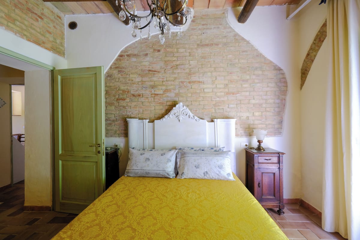 Une chambre avec un mur en briques, une porte en bois, un lit recouvert d’une couverture jaune, une table de chevet avec une lampe et un lustre.