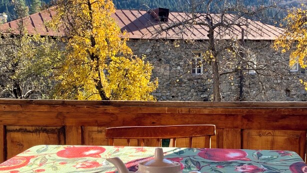 A table with a teapot and cup set on a tablecloth with a fruit pattern, overlooking a mountainous landscape with autumn-colored trees and a stone building with a red roof.