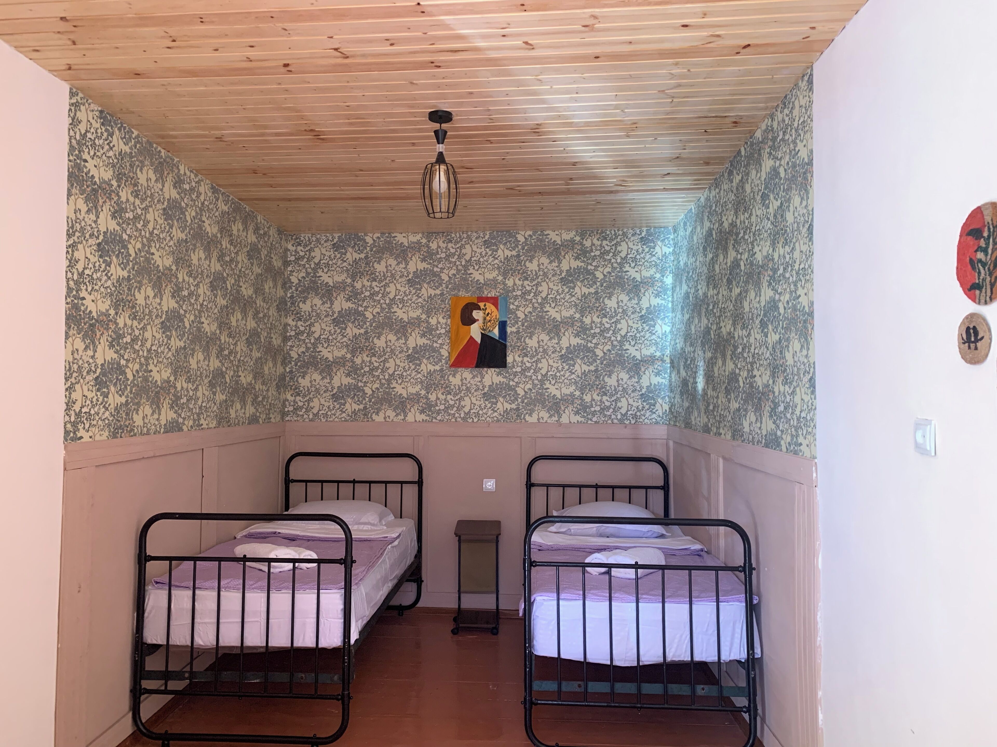 Ein kleines Zimmer mit zwei Betten, Holzboden, Blumenwallpaper und einer einzelnen Lampe.