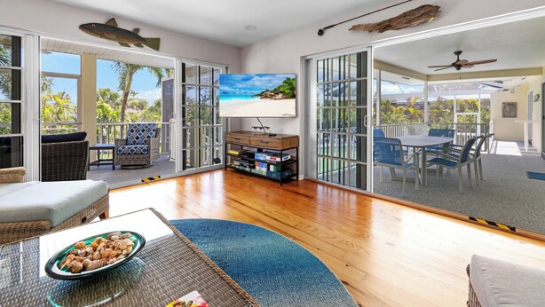 A spacious living area with a large TV, wicker furniture, and a view of the outdoors. - Beach Daze: Island Style Charm on Canal, Quick Walk to Beach! (Sanibel)