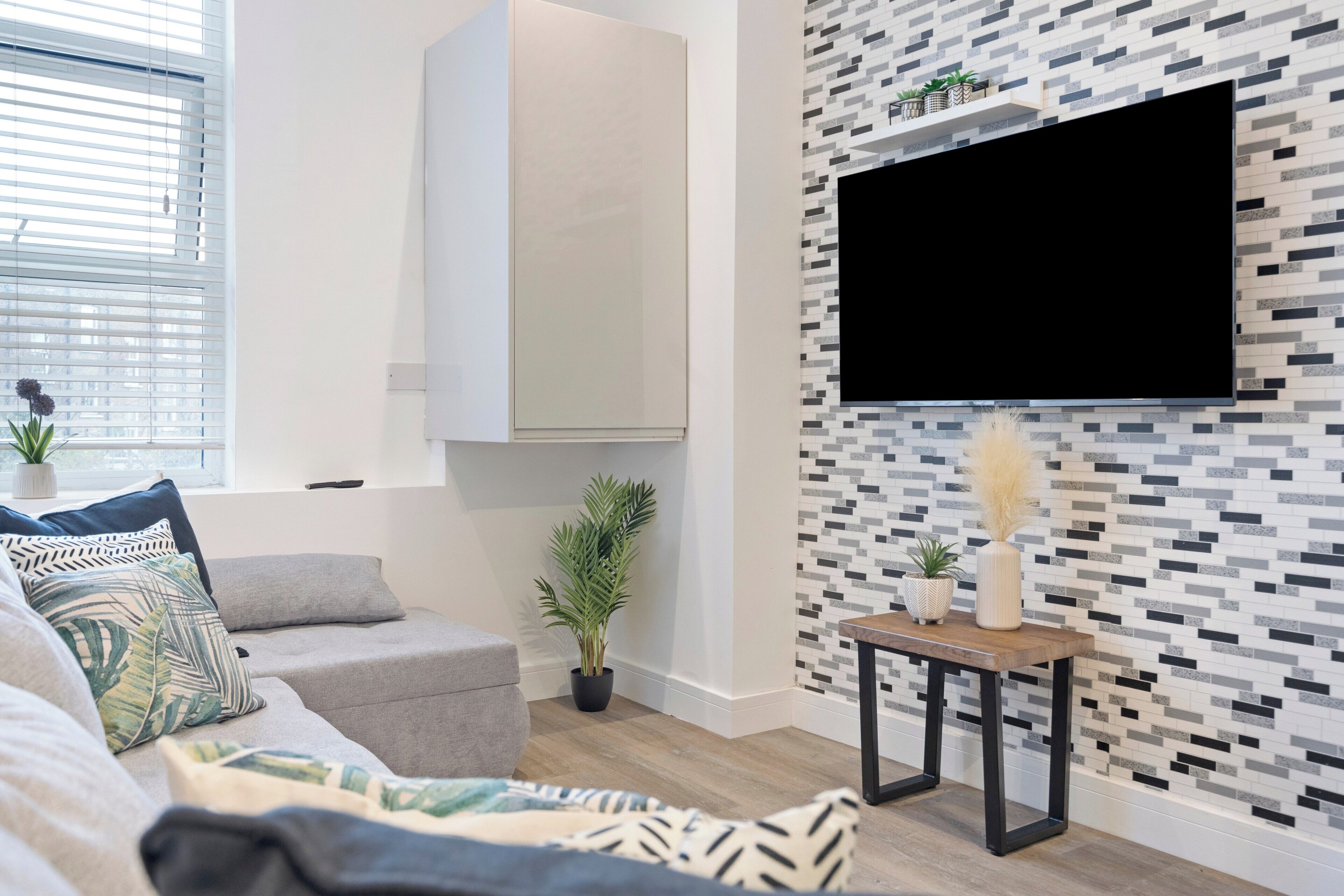 A modern living room with a grey sofa, a mounted TV, a small table with plants, and a patterned wall.
