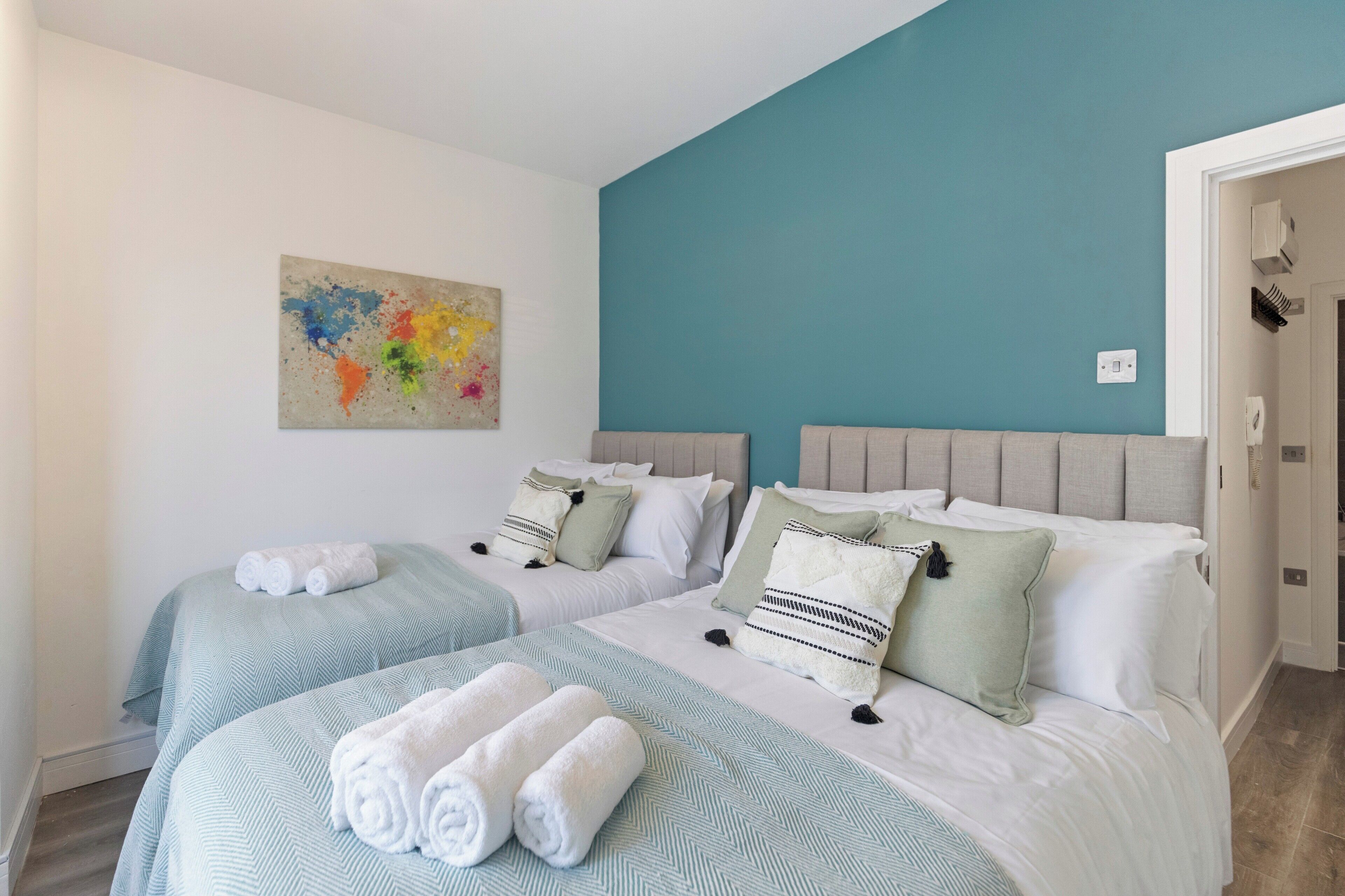 A hotel room with two beds, each with a light blue bedspread and white pillows. There is a teal-colored accent wall and a piece of abstract art on the wall.