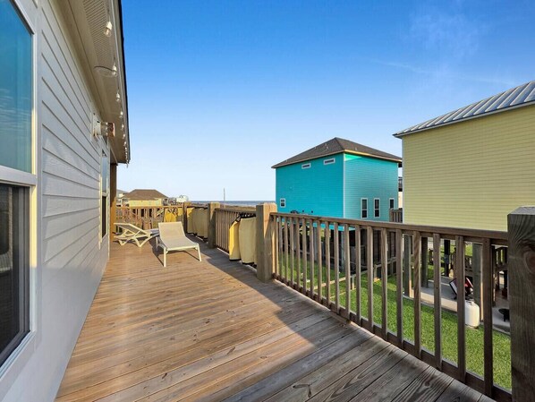 A wooden deck with outdoor furniture, a fenced area, and buildings in the background. - Ocean Views•2 Kings•Sleeps 8•Pet Friendly•Fenced (Bolivar Peninsula)