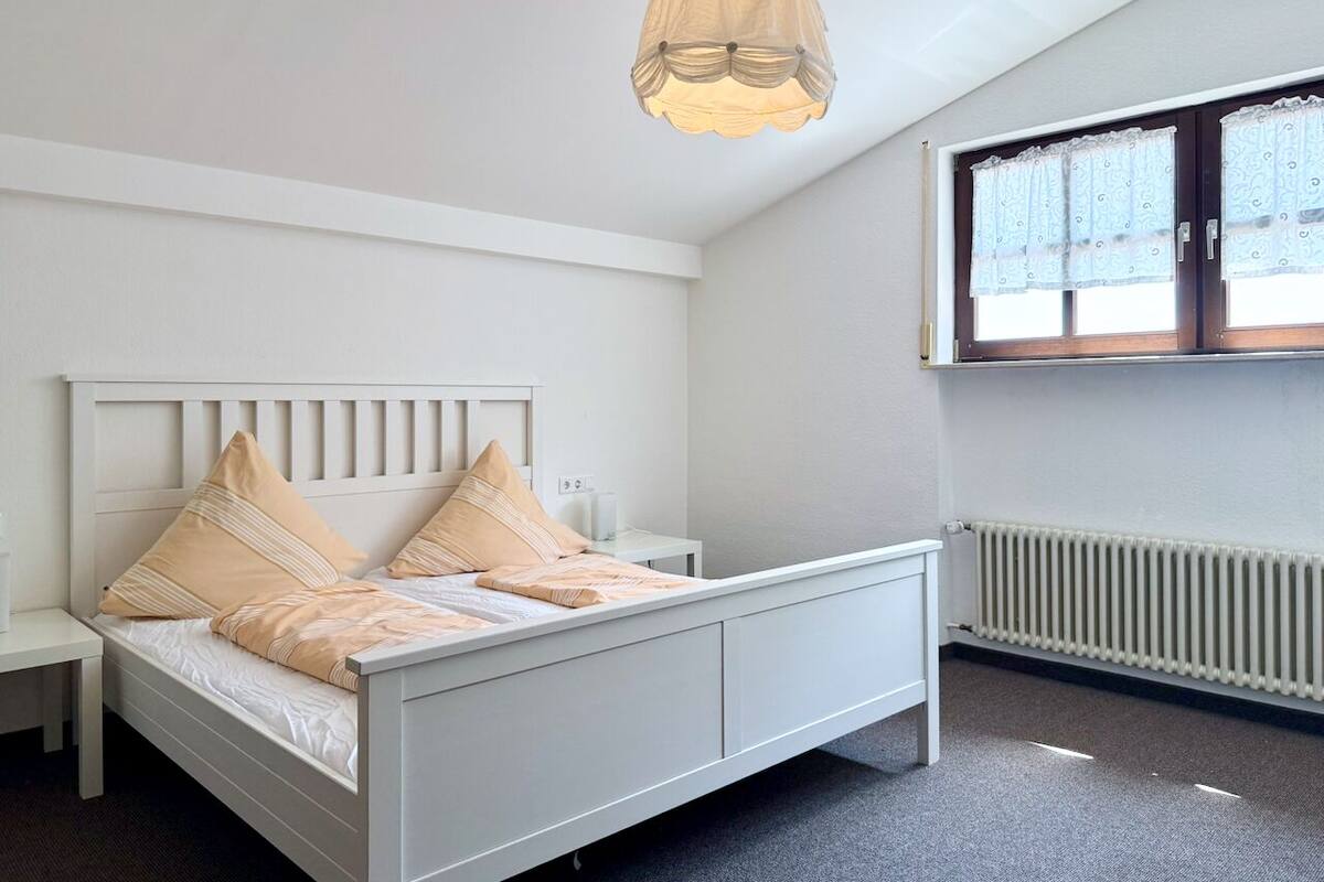 Ein Schlafzimmer mit einem weißen Bett, beige farbenen Kissen, einem Nachttisch, einer Pendelleuchte, einem Fenster mit Vorhängen und einer Heizung.