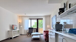 Een moderne woonkamer met een bank, salontafel, tv op een standaard, een fauteuil, en een keukenhoek met een aanrecht en apparatuur.