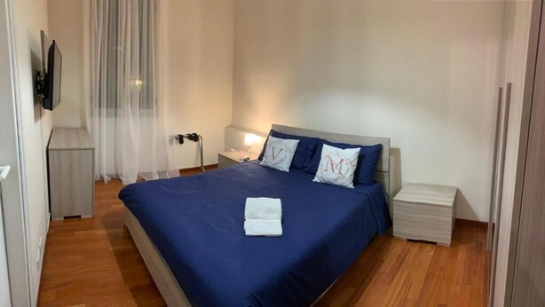 A neatly made bed with a blue bedspread and two pillows, a bedside table with a lamp, and a small nightstand. - Room-charming BnB in the Heart of Ravenn (Ravenna)