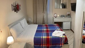 A hotel room with a bed, a bedside table with a lamp, a desk with a chair, a television, and a mirror.