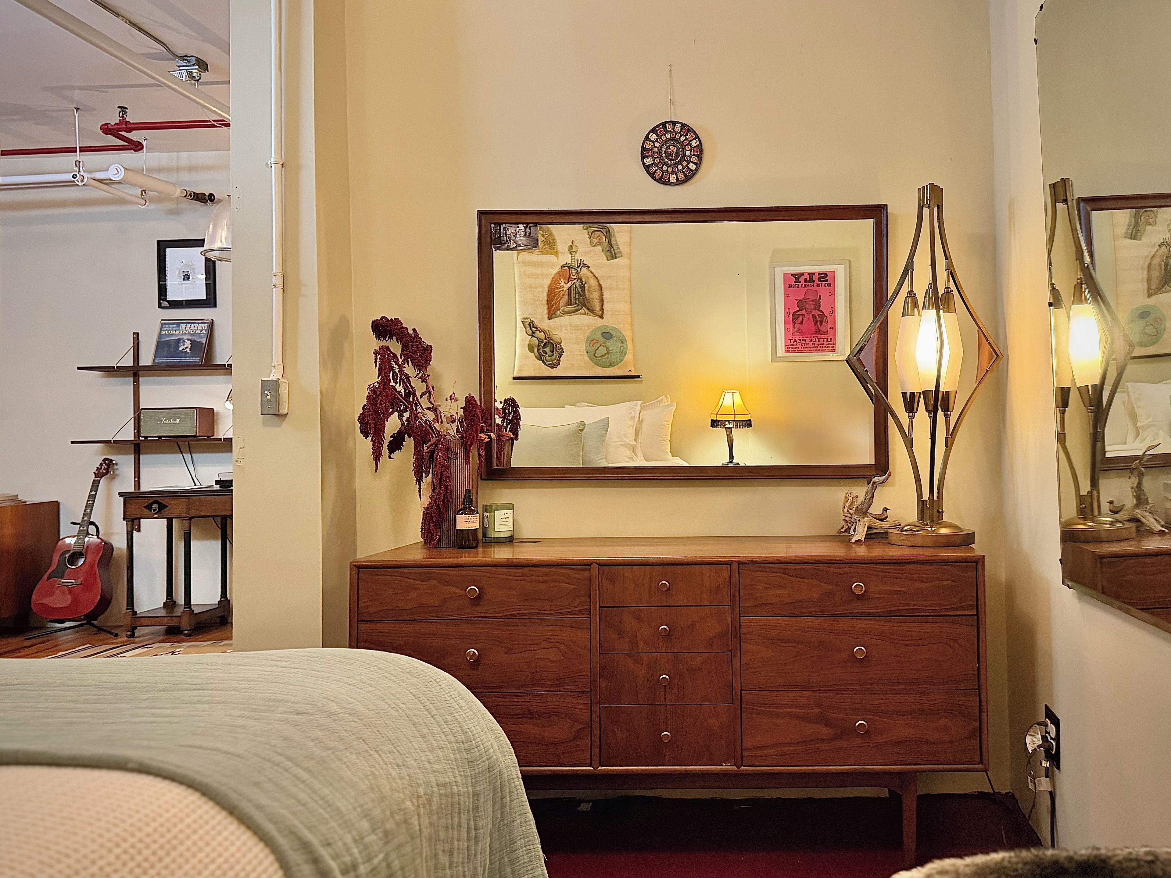 Un dormitorio con un aparador de madera, una cama, una guitarra y un espejo con obras de arte.