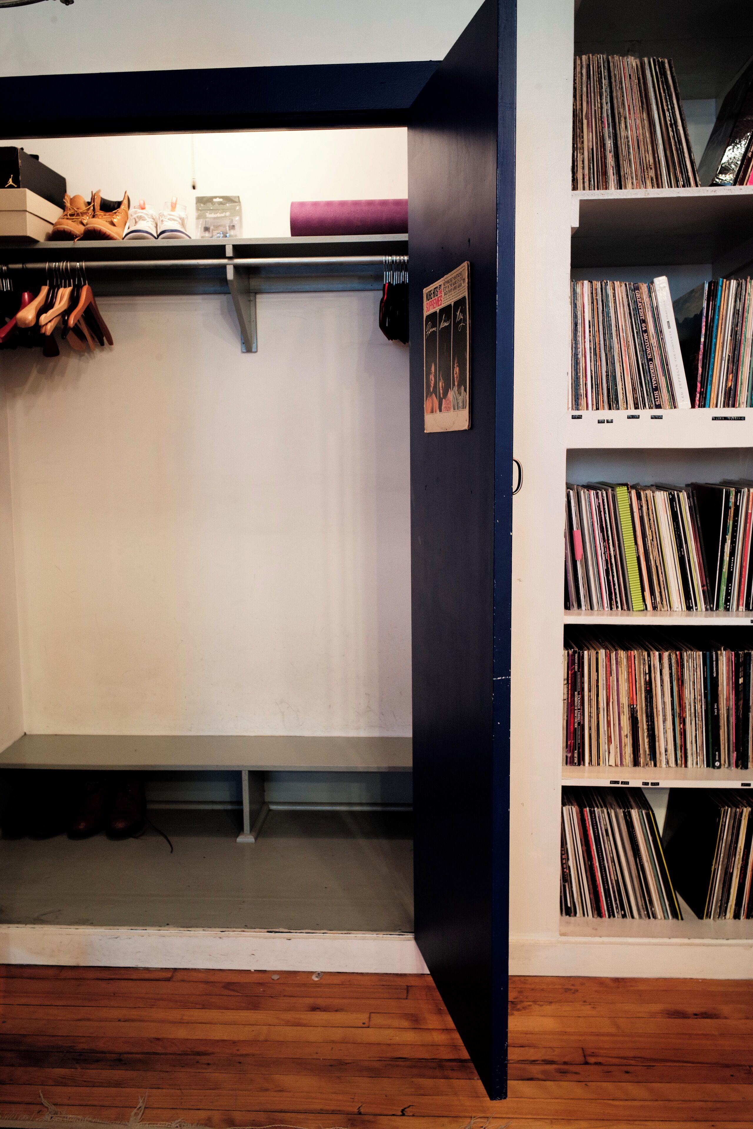 Una habitación con piso de madera, una estantería llena de libros, un zapatero y una puerta que da a otra habitación.