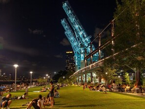 Un parque con gente descansando sobre el césped, un puente y una torre iluminada al fondo.