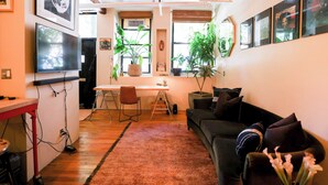 A modern living room with a black sofa, a coffee table, and a TV mounted on the wall. There are several potted plants and framed pictures on the walls. - Bright Artist Loft in Williamsburg Brooklyn (Brooklyn)