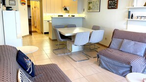 A modern living room with a sofa, a dining table with chairs, a kitchen area, and a hallway leading to another room.