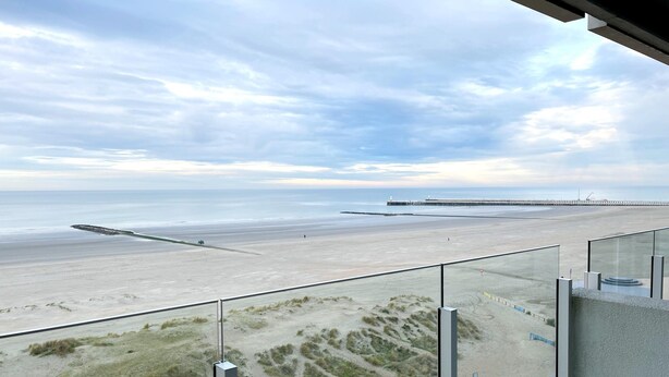 A beach view from a balcony with a glass railing, sandy beach, dunes, and a pier in the distance.