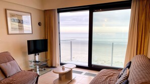 A hotel room with two sofas, a TV, a small table, and a large window with a view of the beach.