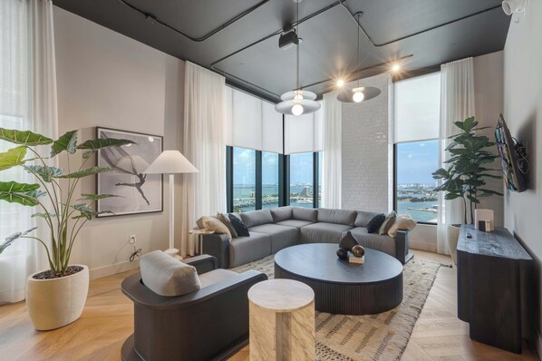 A modern living room with a sectional sofa, round coffee table, and large windows offering a view of the city. - Luxury 1BR | Rooftop Pool | Miami Worldcenter (Miami)