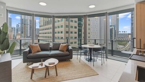 A modern living room with a grey sofa, a small round table, and a large window offering a city view. - Luxury 1BR | Rooftop Pool | Miami Worldcenter (Miami)