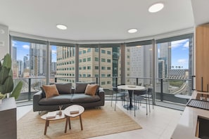 A modern living room with a grey sofa, a small round table, and a large window offering a city view. - Luxury 1BR | Rooftop Pool | Miami Worldcenter (Miami)
