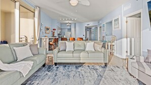A spacious living room with a light-colored sofa, a coffee table, and a ceiling fan. - Shore Thing - Directly on the Beach in Shores! Huge Wrap Around Balcony! (Panama City Beach)