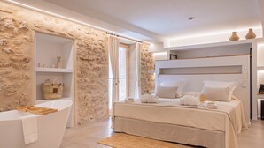 Quarto moderno com parede de pedra, banheira, cama com roupa de cama branca e uma prateleira com objetos decorativos.
