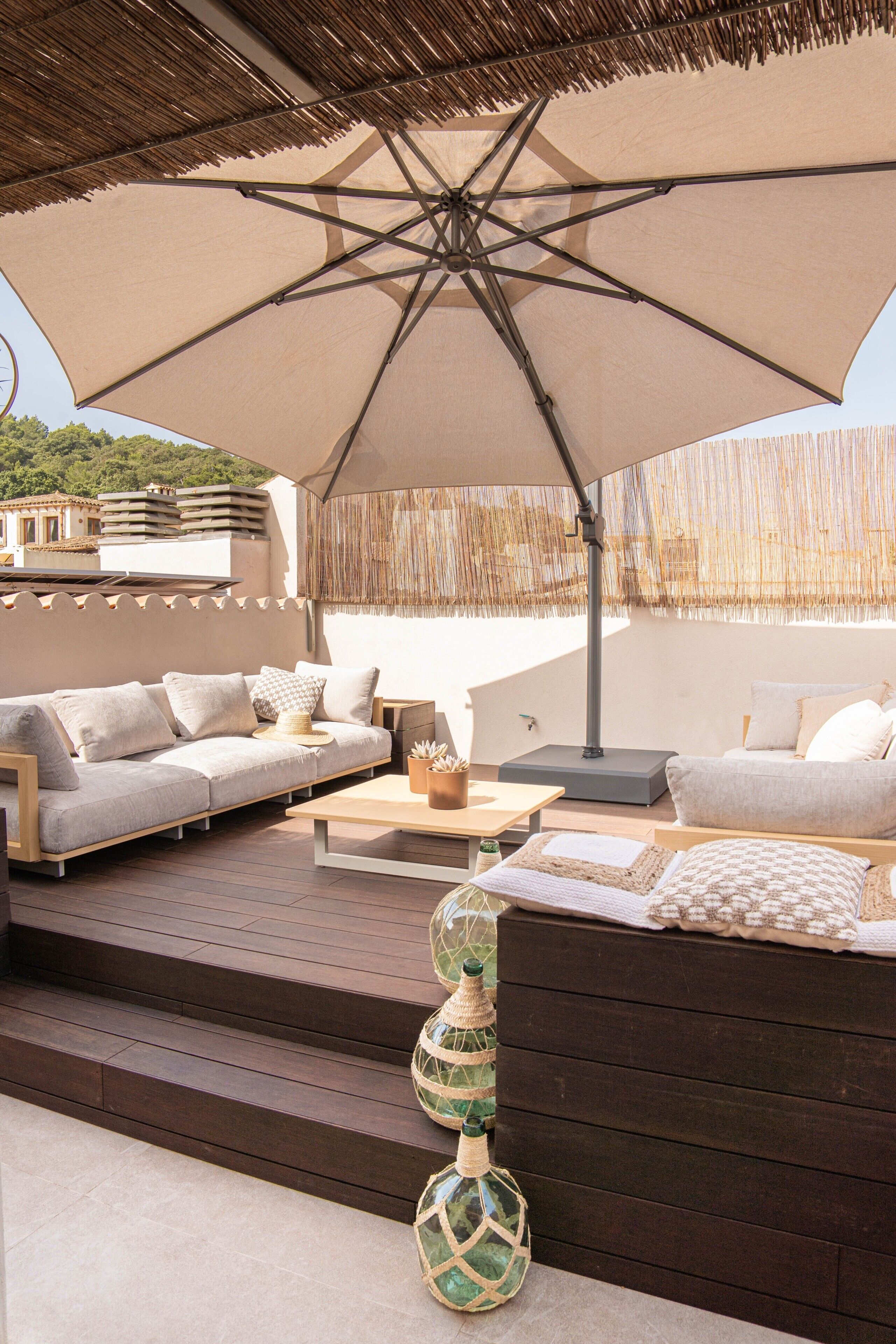 An outdoor seating area with a large umbrella, cushioned sofas, a wooden table, and decorative bottles.