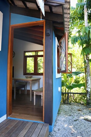 Une porte en bois donne sur une pièce où se trouvent une table et des chaises, une fenêtre et une vue sur la verdure à l’extérieur.