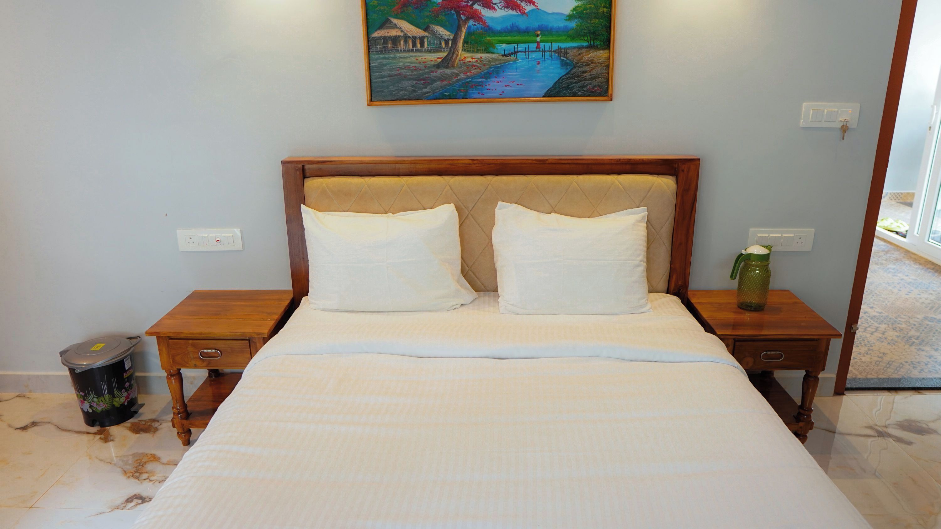 A neatly made bed with a headboard, two bedside tables, a trash bin, and a painting on the wall.