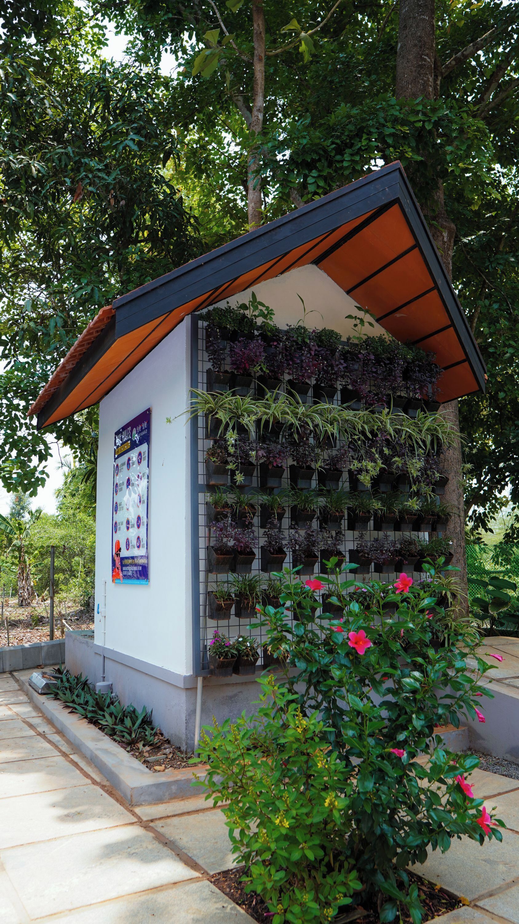 Une petite structure dotée d’un toit incliné, un jardinier mural contenant diverses plantes et un parterre de fleurs roses.