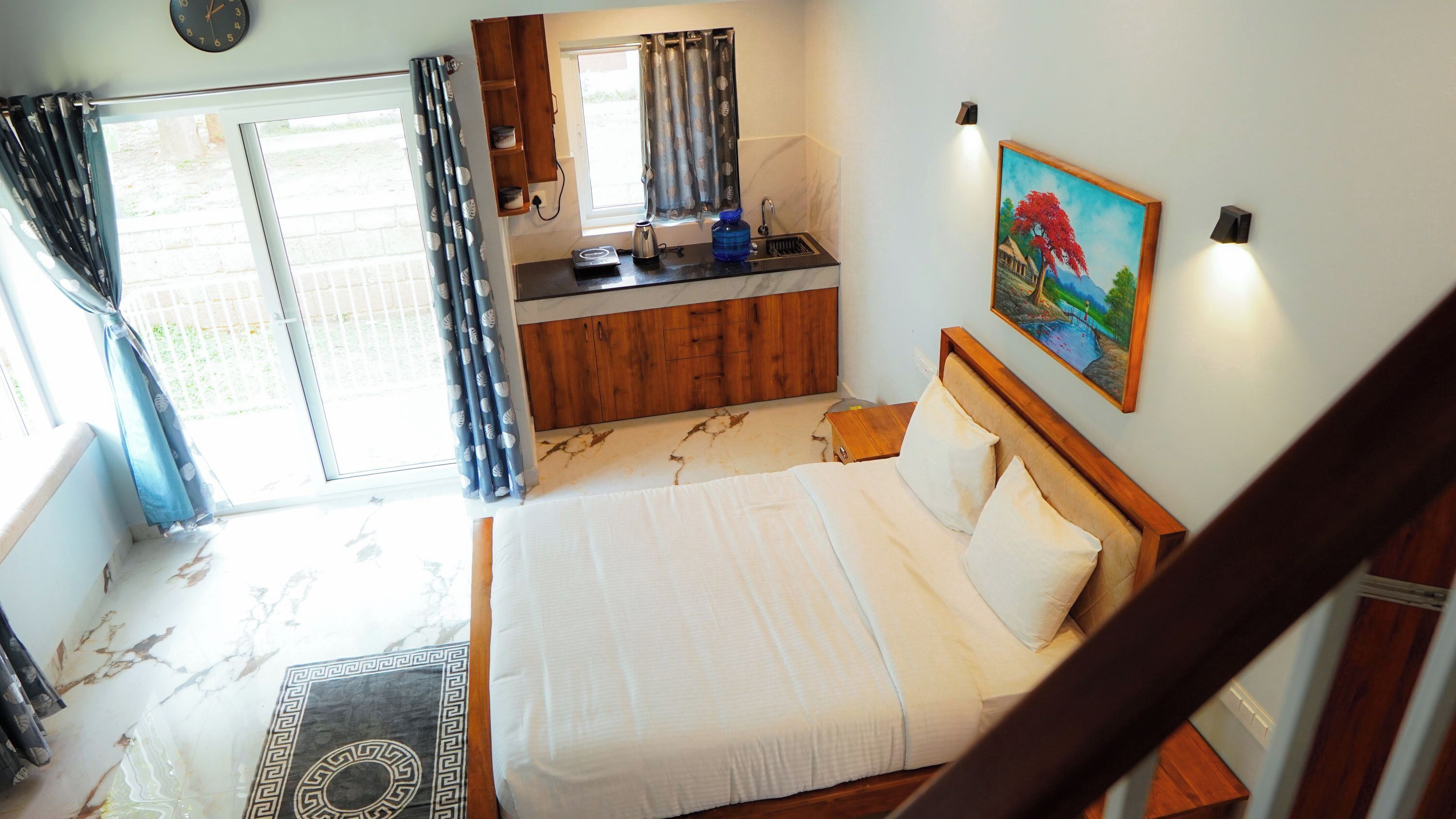 A bedroom with a bed, a small kitchenette, a window with curtains, and a painting on the wall.