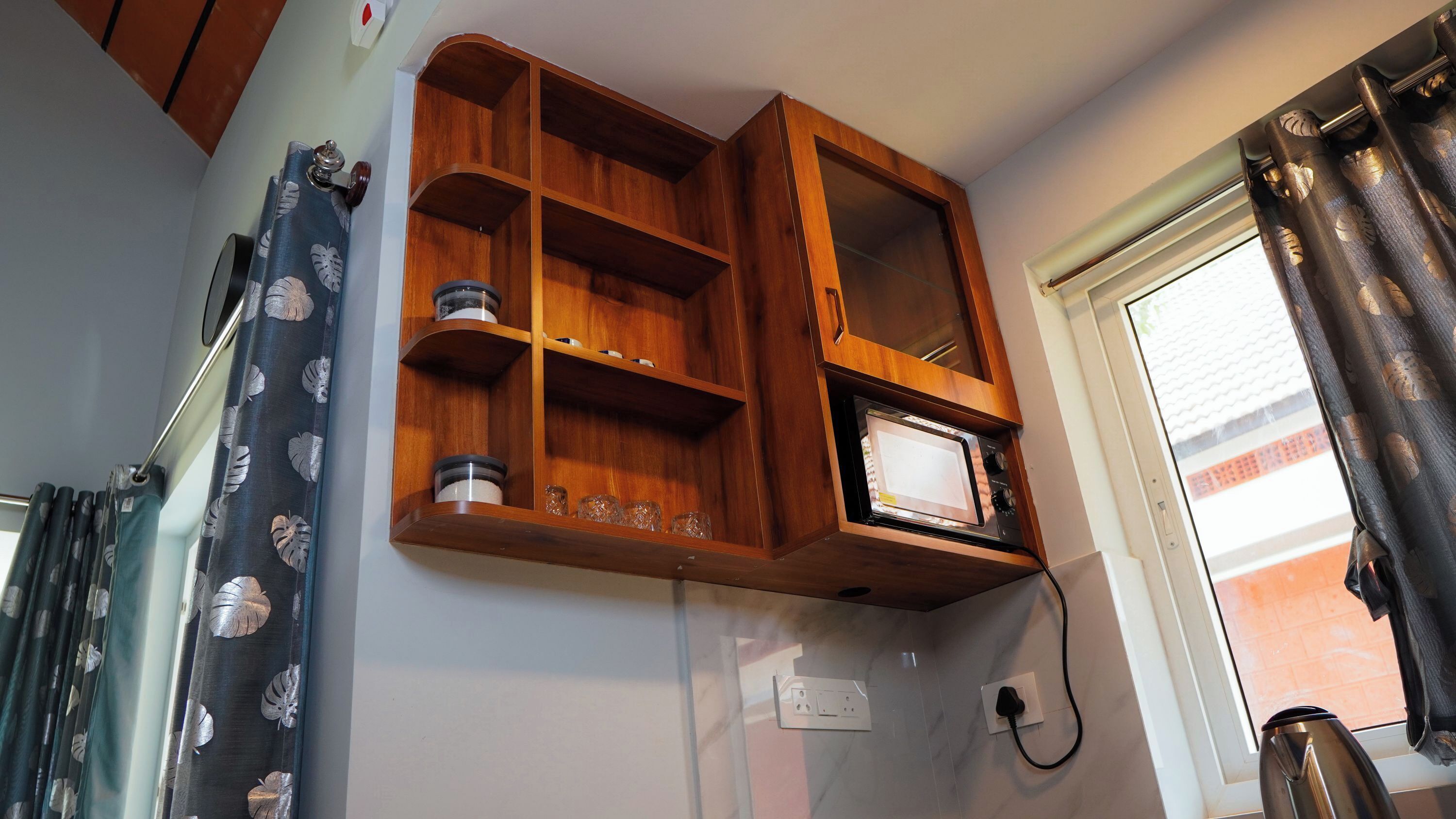 A kitchen with a built-in microwave, wooden shelves, and a window with curtains.