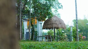 A thatched-roof structure, a bench, and a fenced garden area with trees and plants. - We We Wayanad- Casa de Wayanad (Sulthan Bathery)