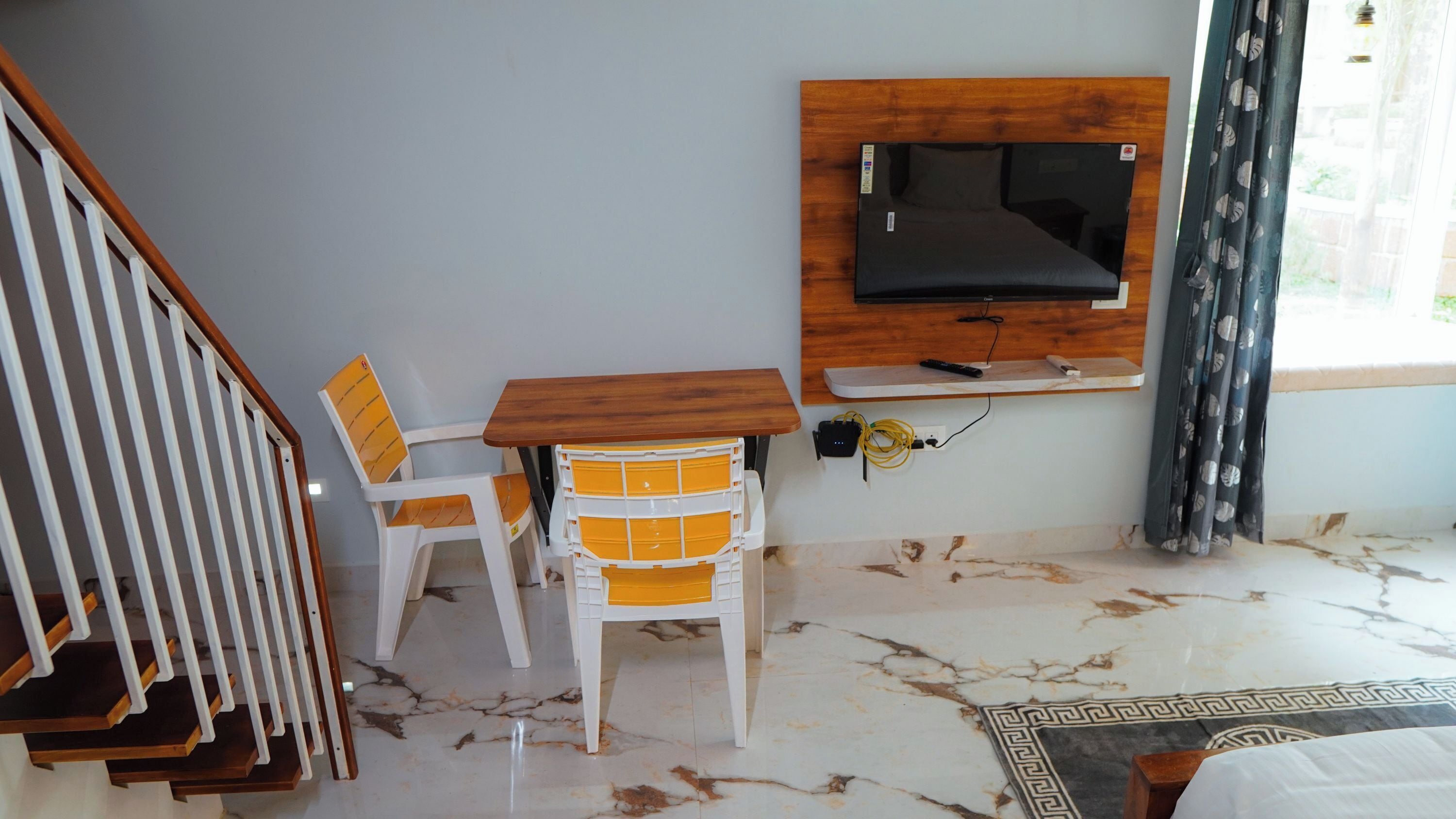 Una habitación con un mueble bajo de madera para el televisor, una silla blanca con cojines amarillos, piso de mármol y una escalera.