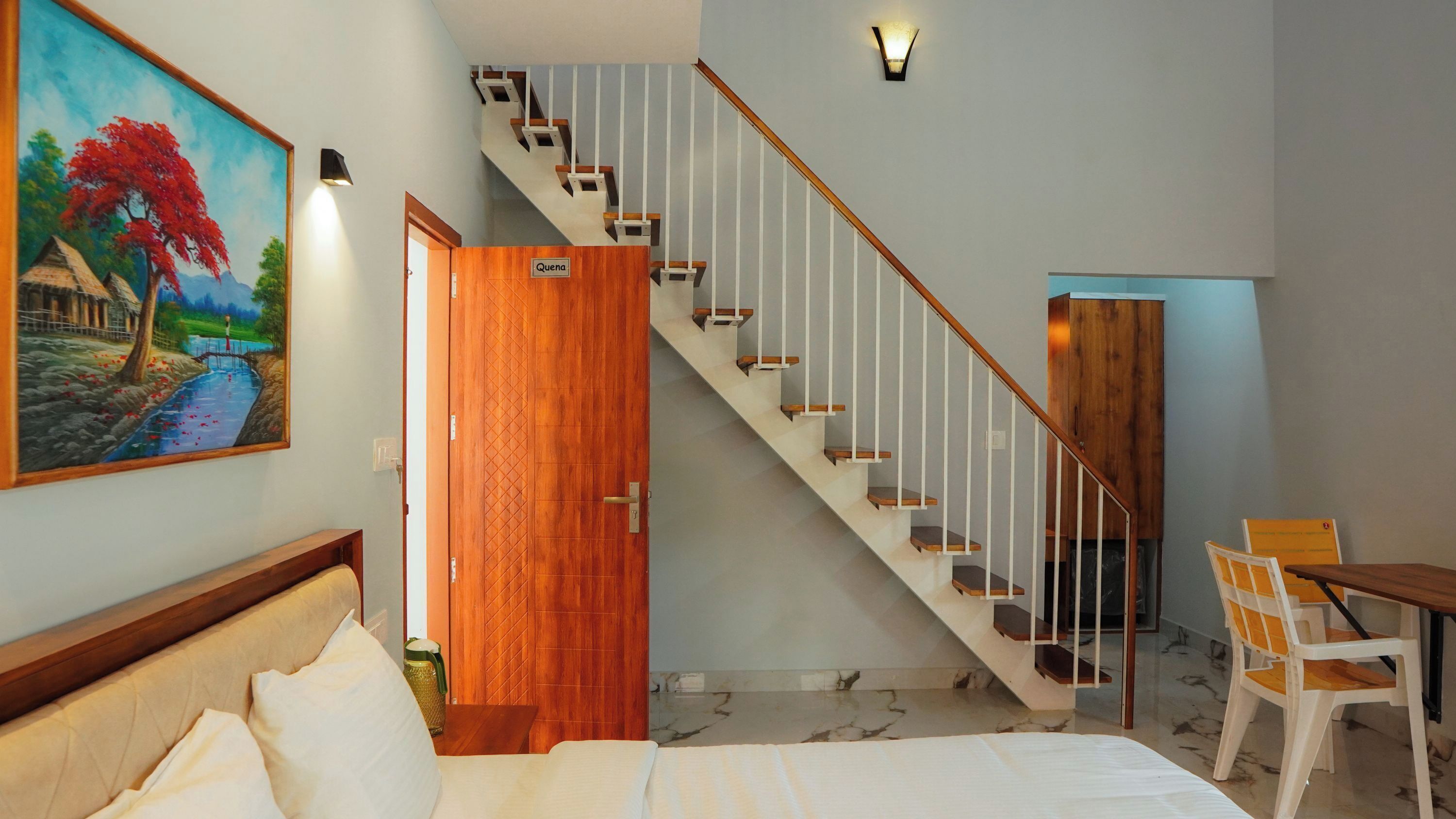 Un dormitorio con cama, escritorio, silla, un cuadro, una escalera y una puerta con la leyenda 'Quena'.