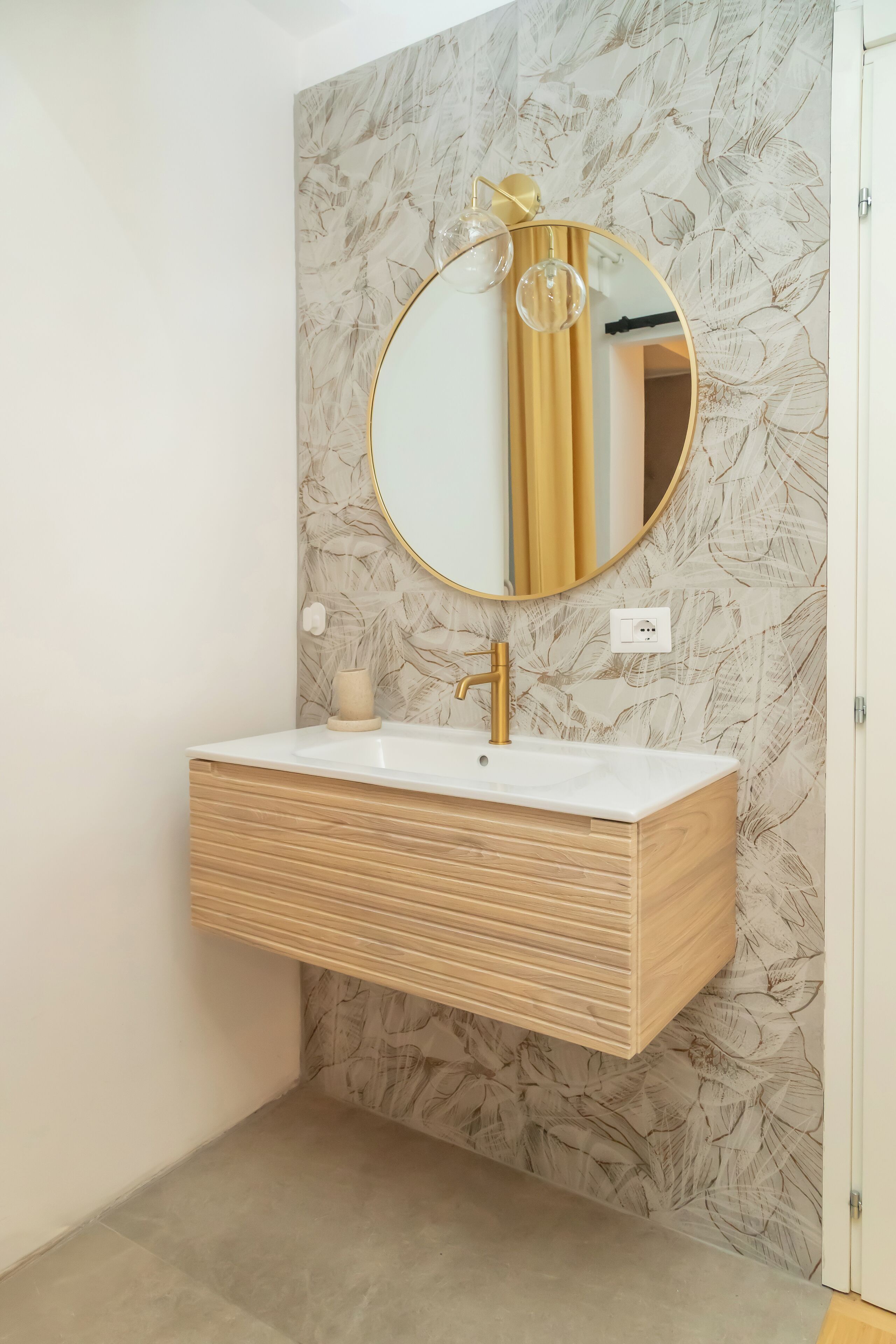 Un baño con pared de diseño de mármol, tocador de madera, grifo dorado y espejo redondo con marco dorado.