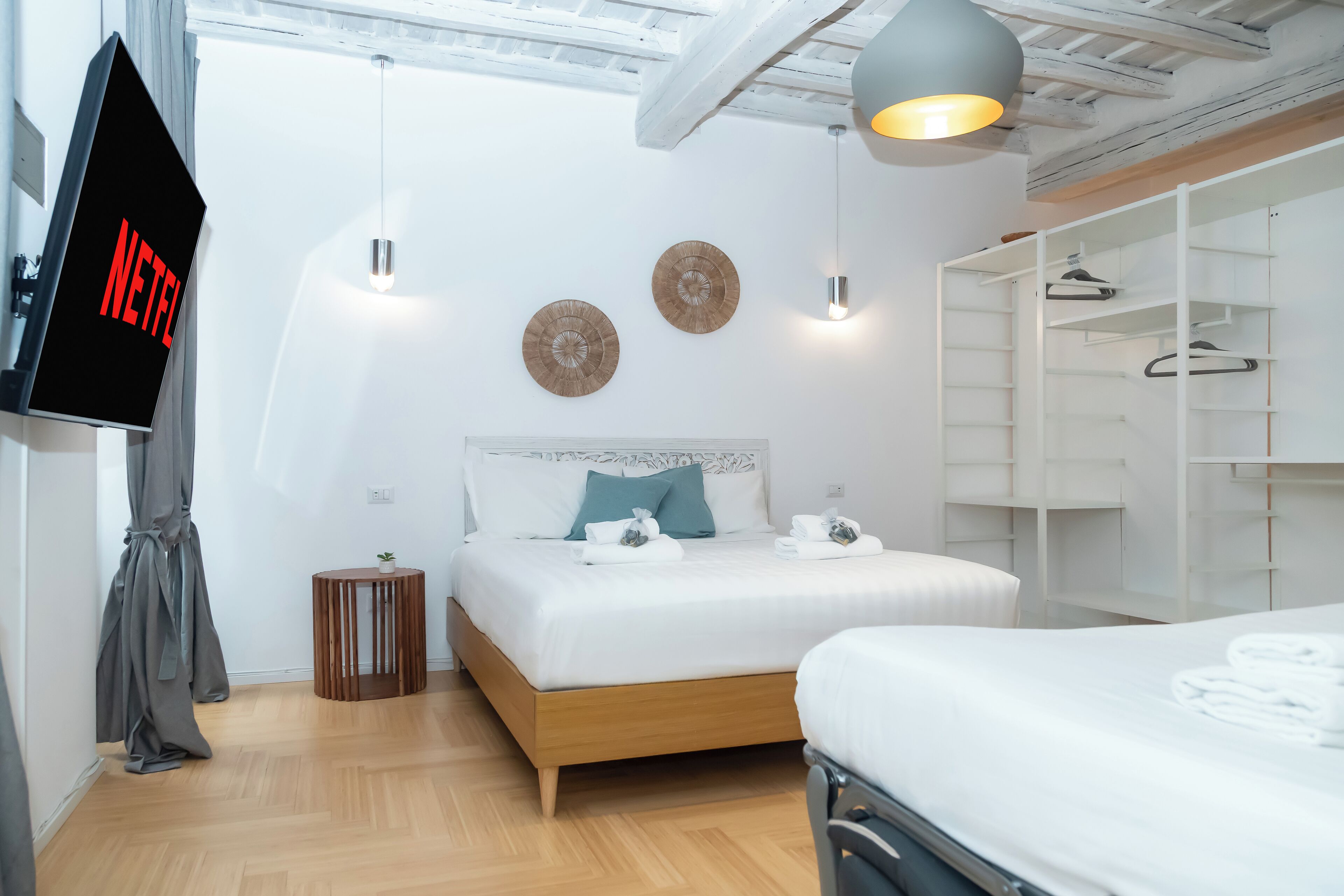 Una habitación de hotel moderna con una cama, una pantalla de televisión mostrando 'NETFLIX', un armario y una pequeña mesa redonda con una planta.