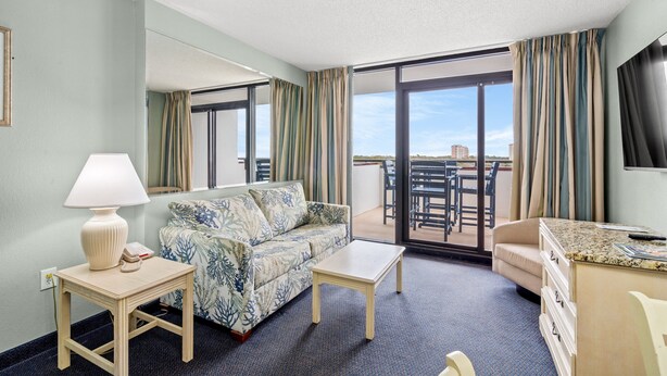 A hotel room with a sofa, a small table with a lamp, a desk, and a TV. The room has a balcony with a view of the city. - Salt life made Simple at Grande Shores (Myrtle Beach)