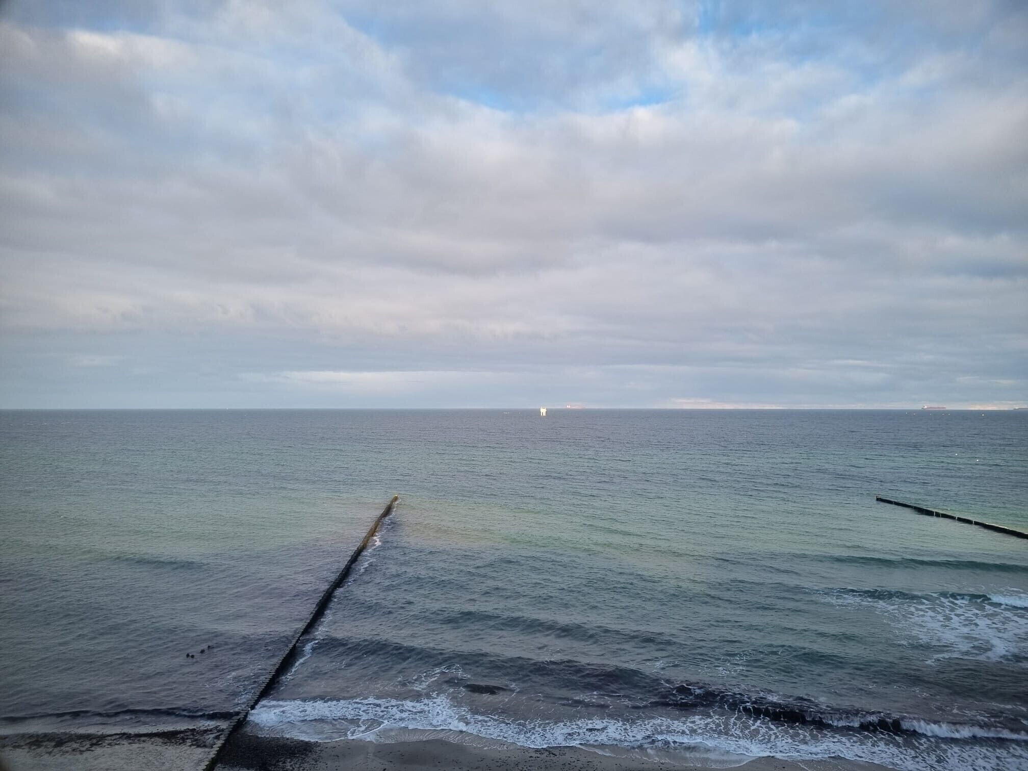 A coastal view with a calm sea, a wooden jetty, and distant ships.