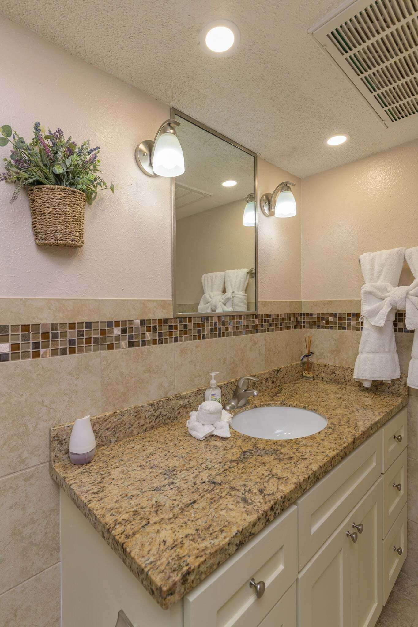 A bathroom with a granite countertop, white sink, and a mirror above it. There is a towel rack with a white towel and a potted plant on the wall.