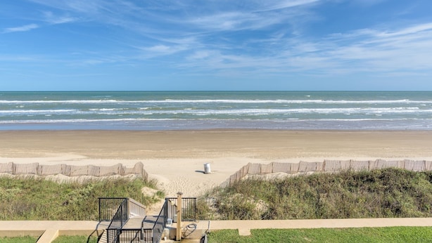 Ein Strand mit Holzzaun, einem Betonweg und einer Rasenfläche, die zum Ufer führt.