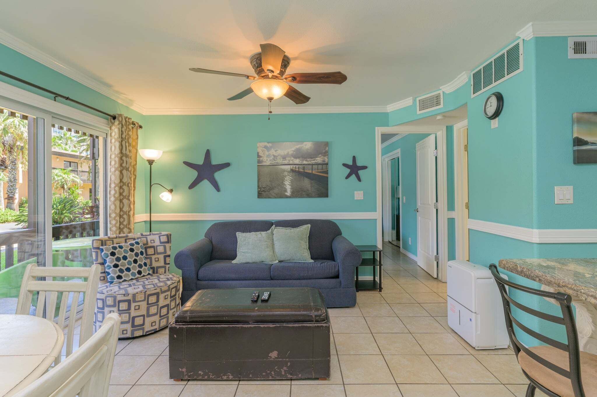 Un salon aux murs de couleur turquoise, doté d’un ventilateur de plafond, d’un canapé, d’une table basse et d’un coin repas avec une table et des chaises.