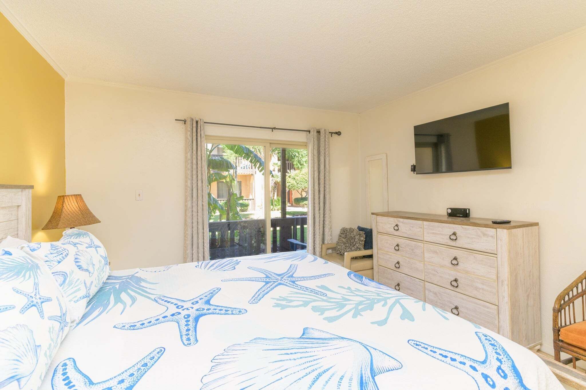 Un dormitorio con cama, cómoda, televisor y balcón con cortinas.