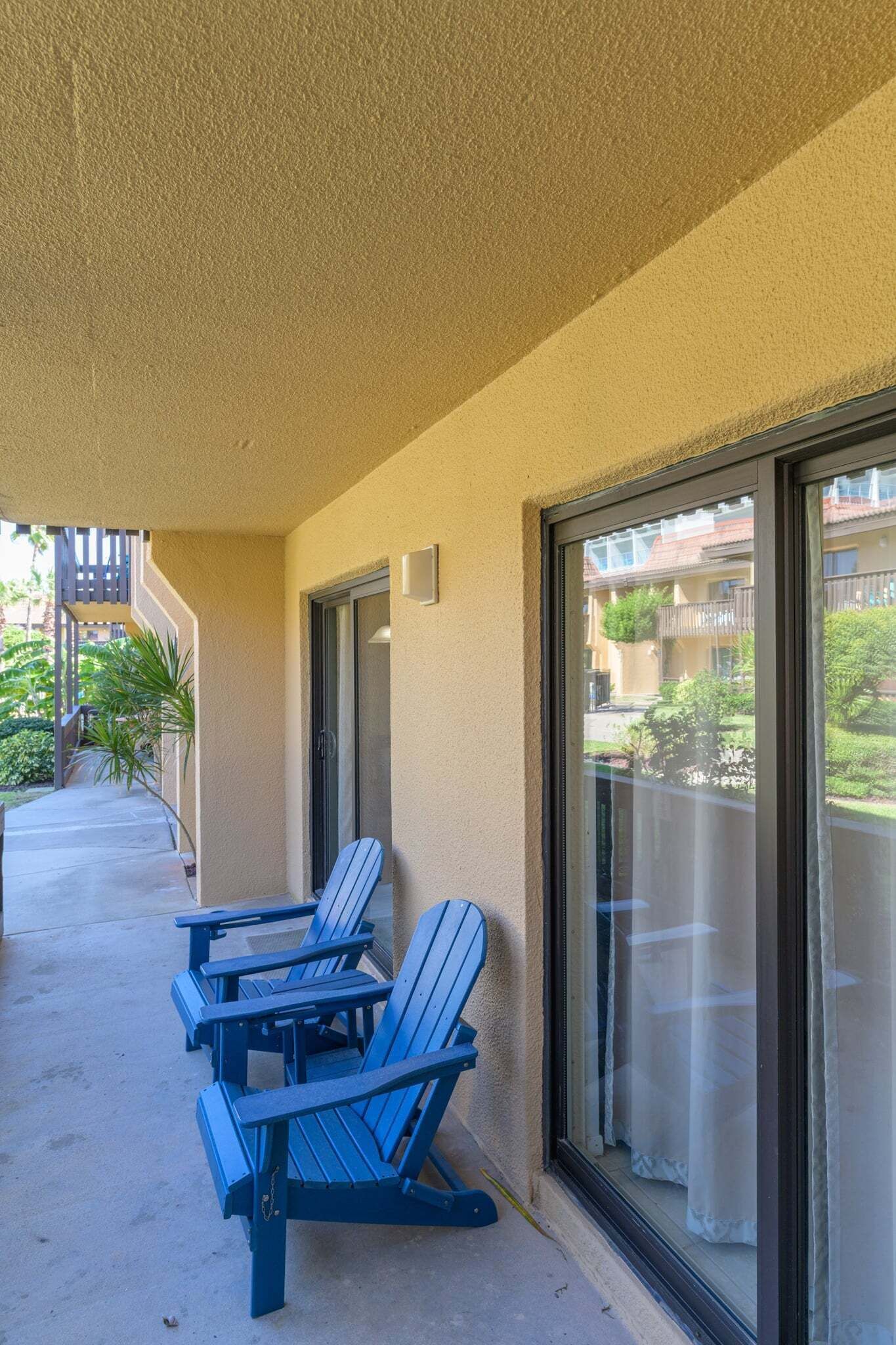 A balcony with two blue Adirondack chairs, a glass door, and a view of a residential area with balconies.