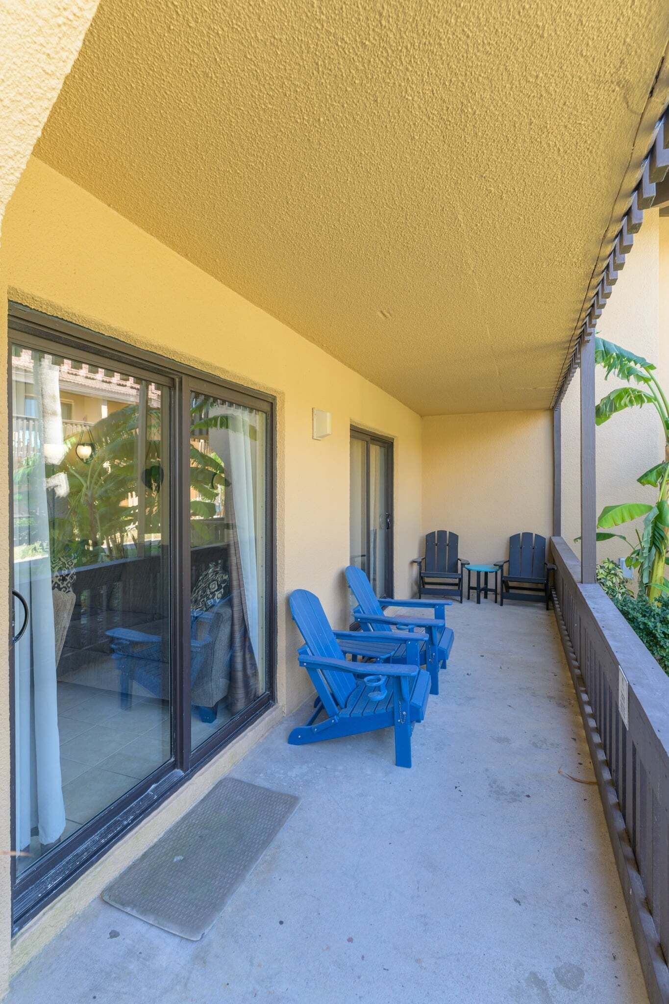 Varanda com cadeiras azuis e uma mesa, porta de correr de vidro e vista para área verde.