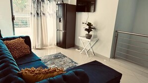Una sala moderna con un sofá azul, una pequeña mesita blanca con una planta, un refrigerador negro y un televisor montado en la pared.