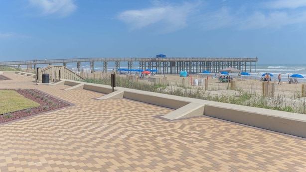 Un paseo marítimo en la playa con un muelle que se adentra en el océano, con un área de descanso cubierta y una escalera que baja a la playa.