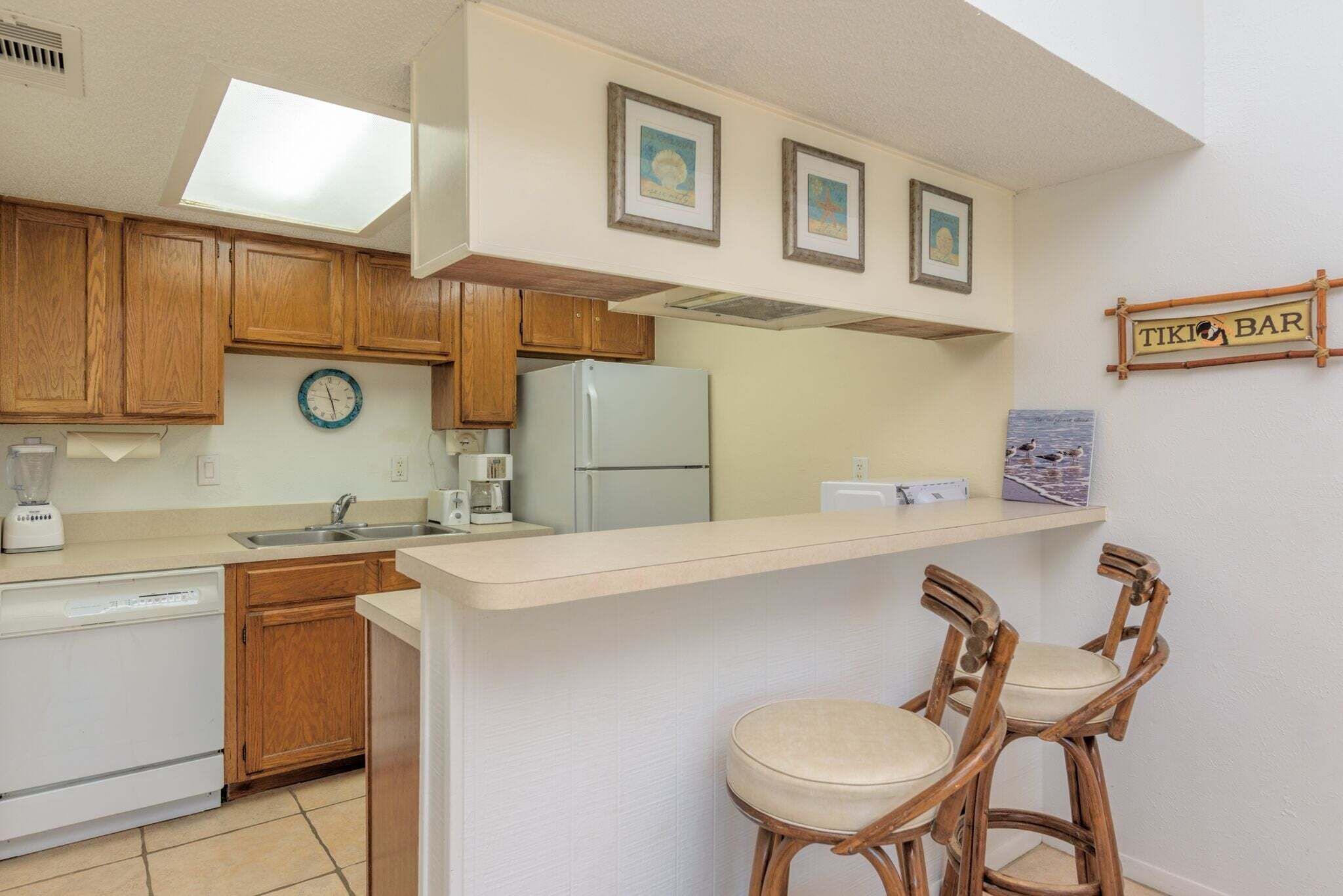 A kitchen with wooden cabinets, a white refrigerator, a coffee maker, and a clock. There are two bar stools and a sign that reads "TIKI BAR".